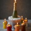 Bougies artisanales en forme de bouteille de champagne et de bouchons colorés, tous allumés et disposés sur un support en marbre avec un fond gris neutre.